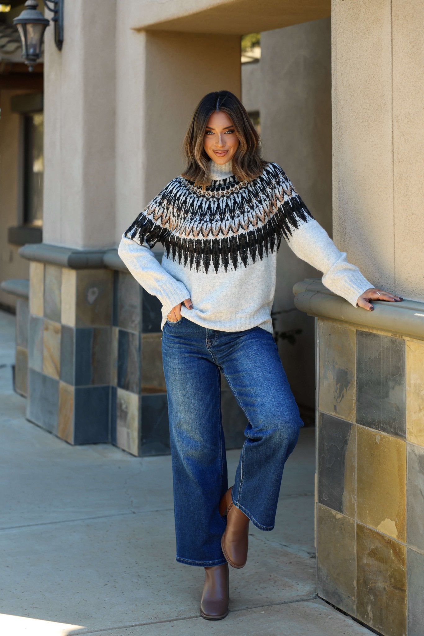 Woman wears the Oatmeal Fair Isle Mock Neck Sweater and wide-leg jeans, smiling as she leans against a pillar with legs crossed.