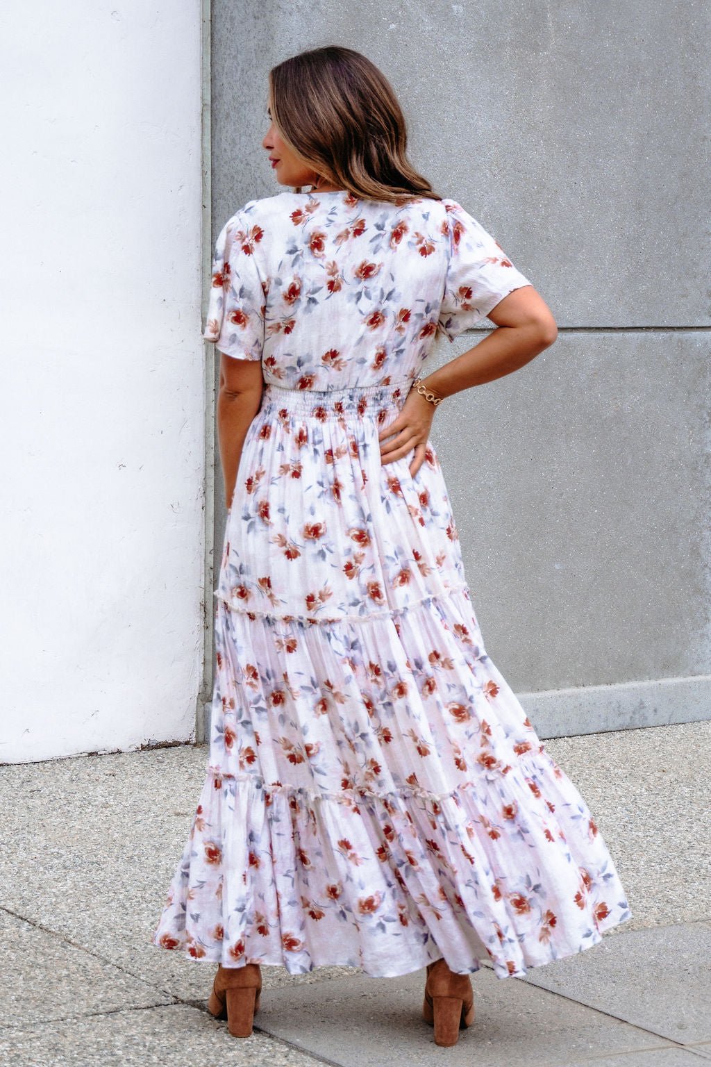 A woman in the Oatmeal Floral Print Smock Tiered Maxi Dress poses outdoors with breezy elegance, standing near a concrete wall.