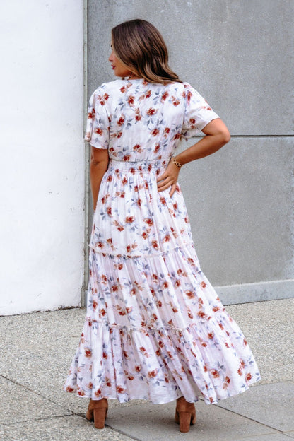 A woman in the Oatmeal Floral Print Smock Tiered Maxi Dress poses outdoors with breezy elegance, standing near a concrete wall.