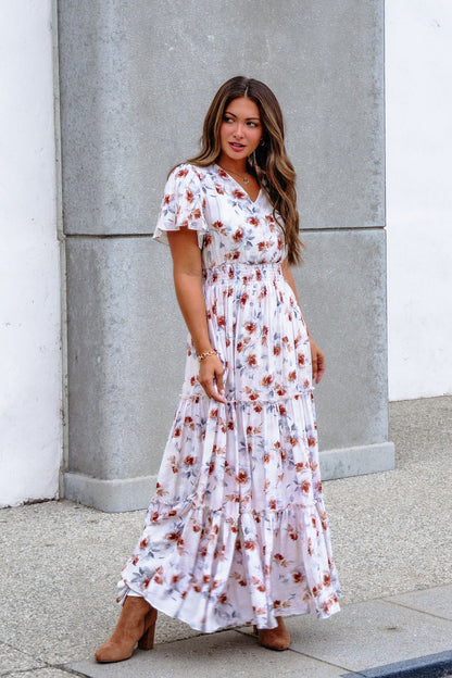 A woman stands outdoors by a concrete wall in the Oatmeal Floral Print Smock Tiered Maxi Dress and brown boots.