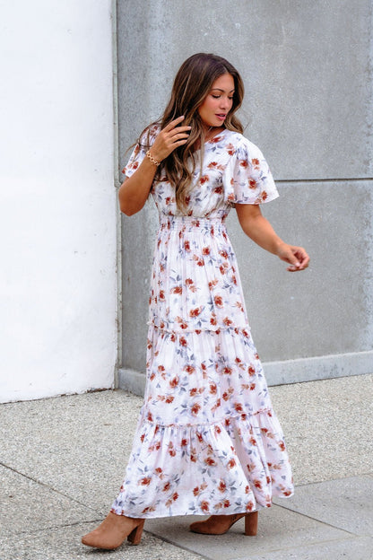 Woman wearing the Oatmeal Floral Print Smock Tiered Maxi Dress walks outdoors, touching her hair by a neutral wall.