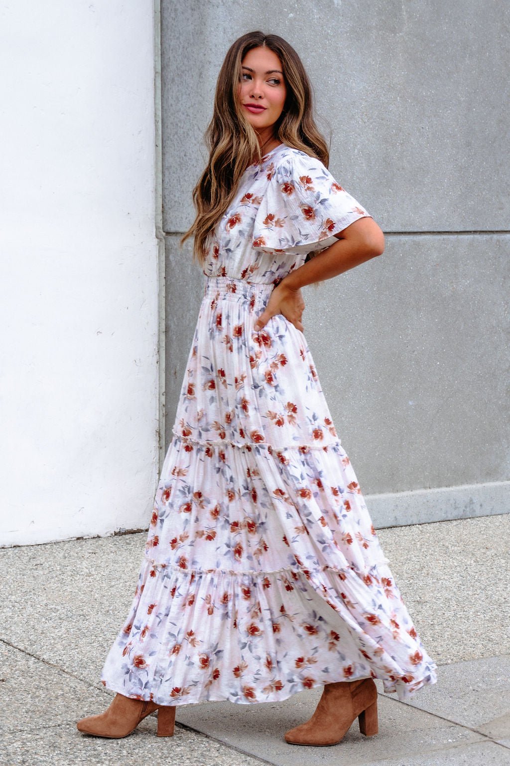 Woman in an Oatmeal Floral Print Smock Tiered Maxi Dress poses sideways by a concrete and white wall.