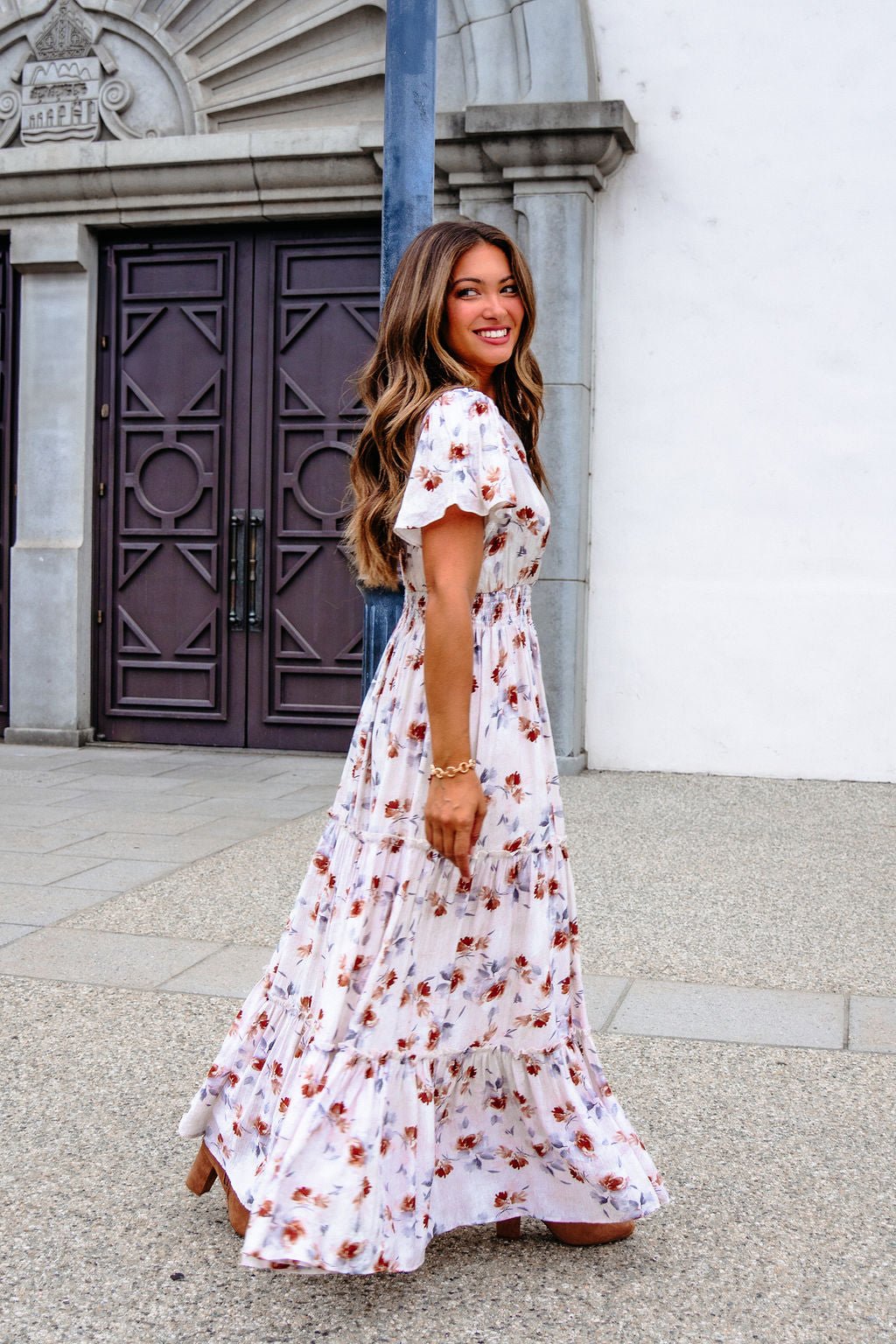 A woman in the Oatmeal Floral Print Smock Tiered Maxi Dress smiles outside a white stone building, radiating breezy elegance.