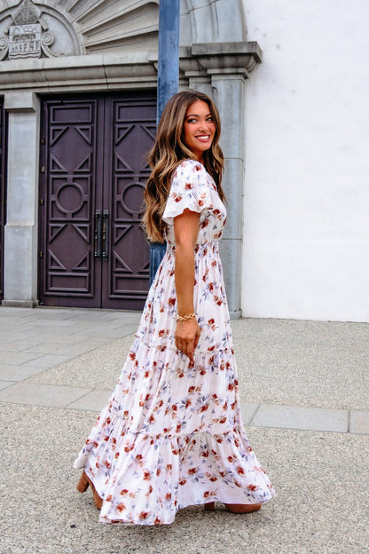 A woman in the Oatmeal Floral Print Smock Tiered Maxi Dress smiles outside a white stone building, radiating breezy elegance.