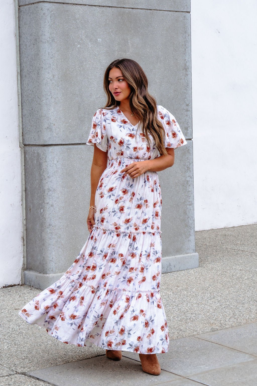Woman in an Oatmeal Floral Print Smock Tiered Maxi Dress stands by a gray stone wall, looking to the side.
