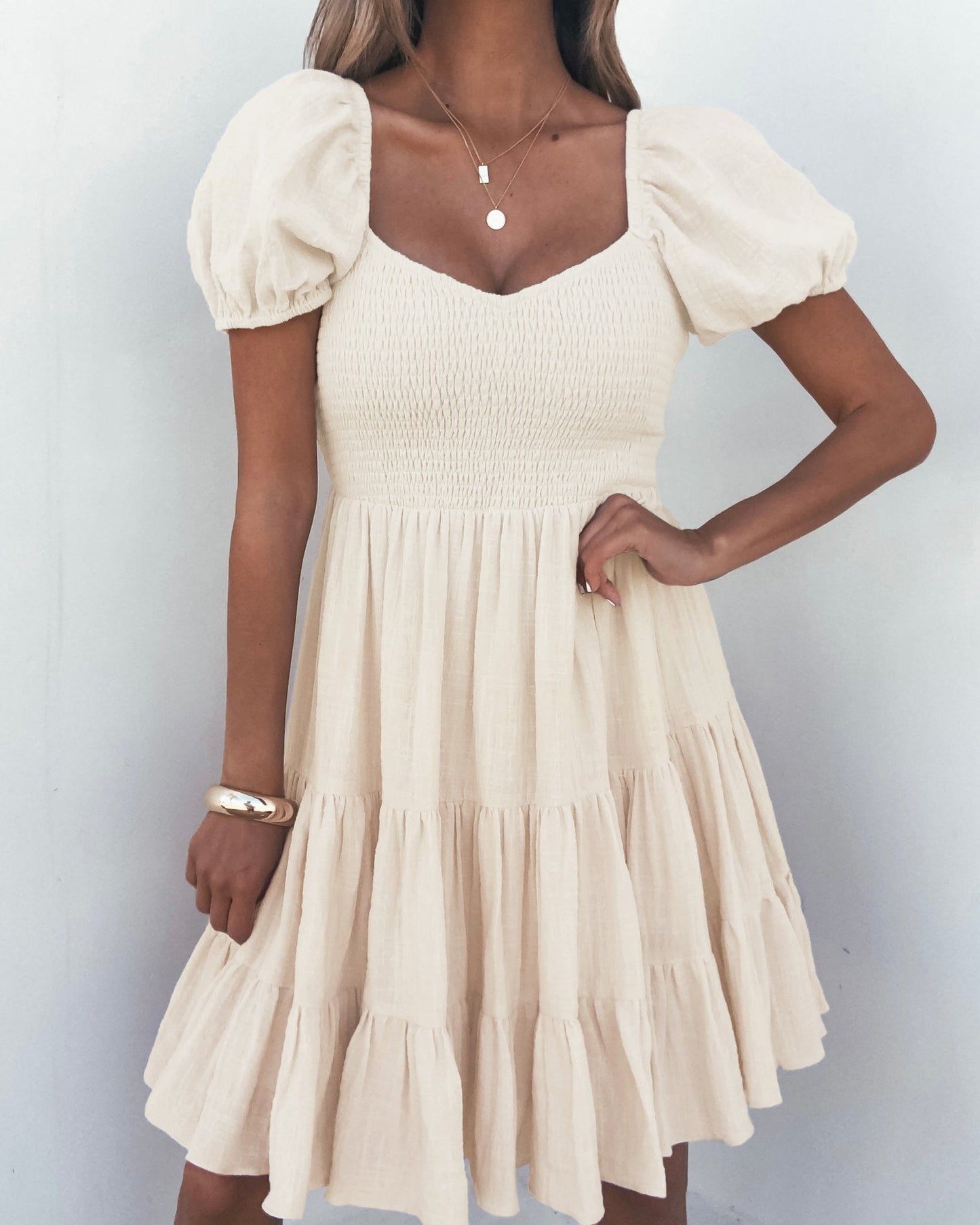A woman wears the Oatmeal Puff Sleeve Tiered Mini Dress - FINAL SALE with layered necklaces and a bracelet against a white wall.