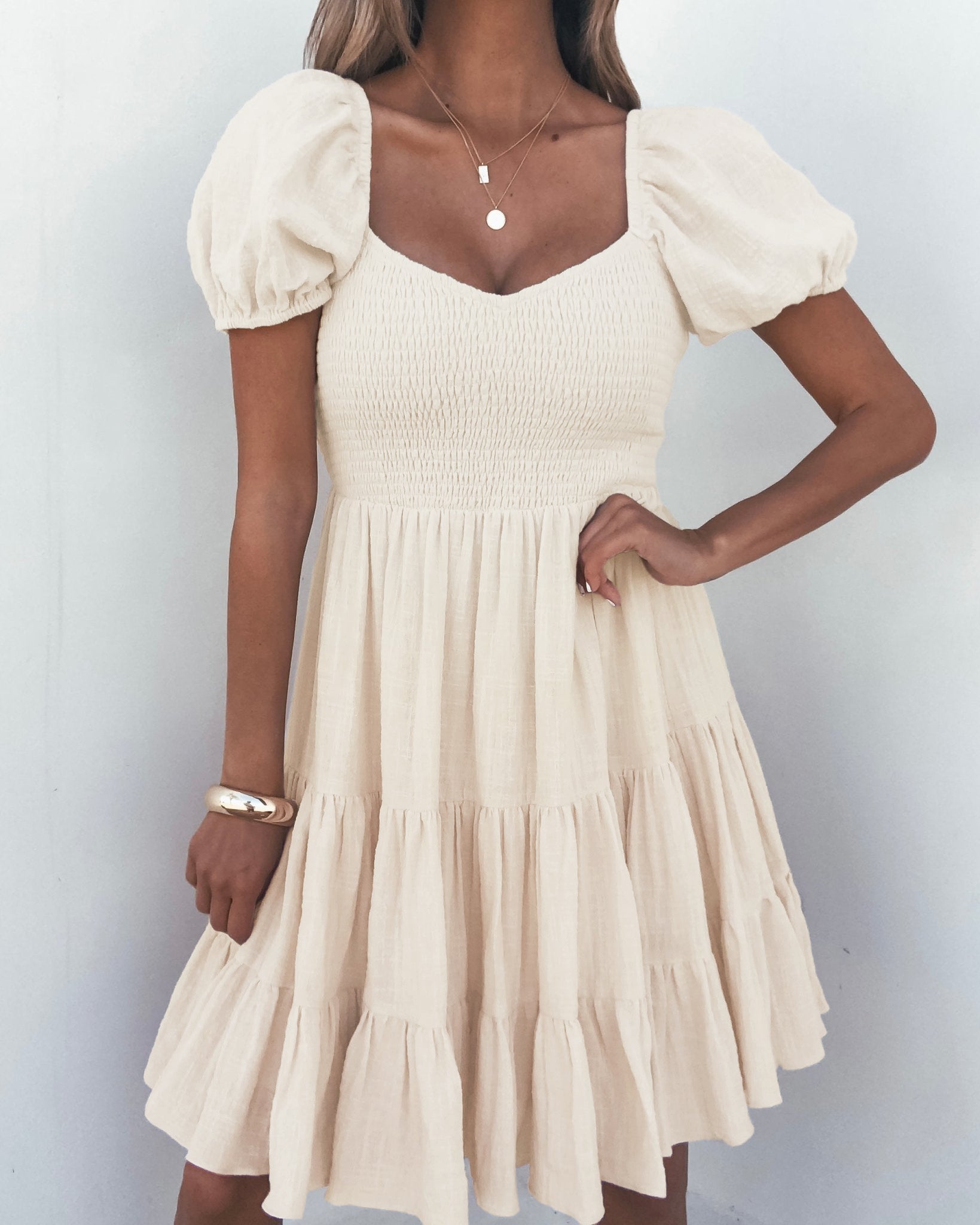A woman wears the Oatmeal Puff Sleeve Tiered Mini Dress - FINAL SALE with layered necklaces and a bracelet against a white wall.
