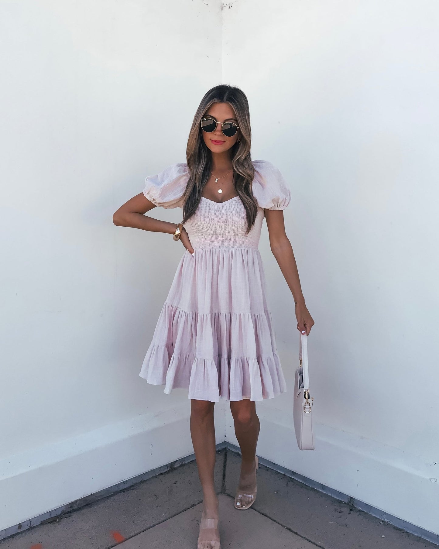 Woman in an Oatmeal Puff Sleeve Tiered Mini Dress, nude sandals, sunglasses, and a white bag stands against a white wall.