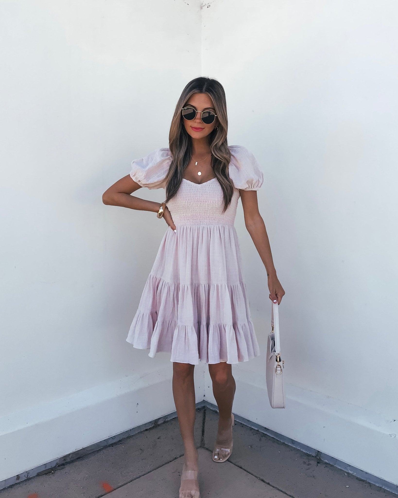 Woman in an Oatmeal Puff Sleeve Tiered Mini Dress, nude sandals, sunglasses, and a white bag stands against a white wall.