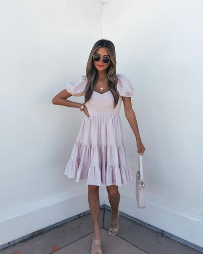 Woman in an Oatmeal Puff Sleeve Tiered Mini Dress, nude sandals, sunglasses, and a white bag stands against a white wall.