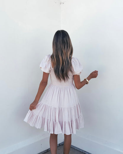 Woman with long, wavy hair wears an Oatmeal Puff Sleeve Tiered Mini Dress, holding the skirt as she faces a white corner.