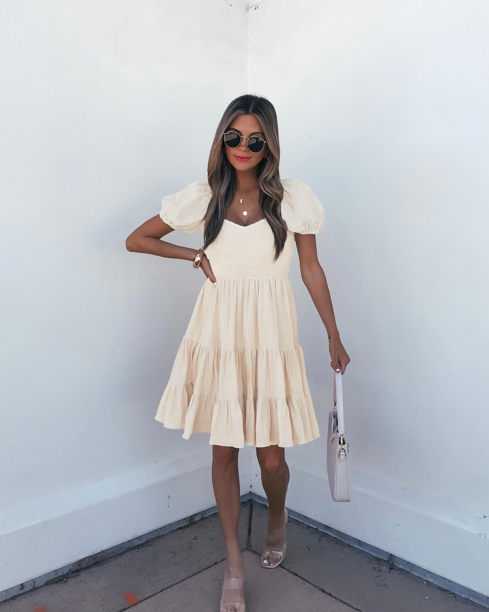 Woman in an Oatmeal Puff Sleeve Tiered Mini Dress - FINAL SALE with sunglasses, sandals, and a white bag stands by a white wall.