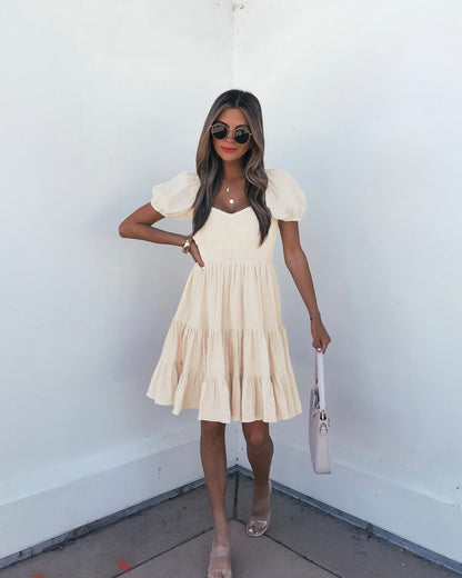 Woman in an Oatmeal Puff Sleeve Tiered Mini Dress - FINAL SALE with sunglasses, sandals, and a white bag stands by a white wall.