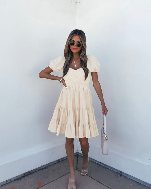 Woman in an Oatmeal Puff Sleeve Tiered Mini Dress, sunglasses, sandals, holding a white handbag by a white corner wall.