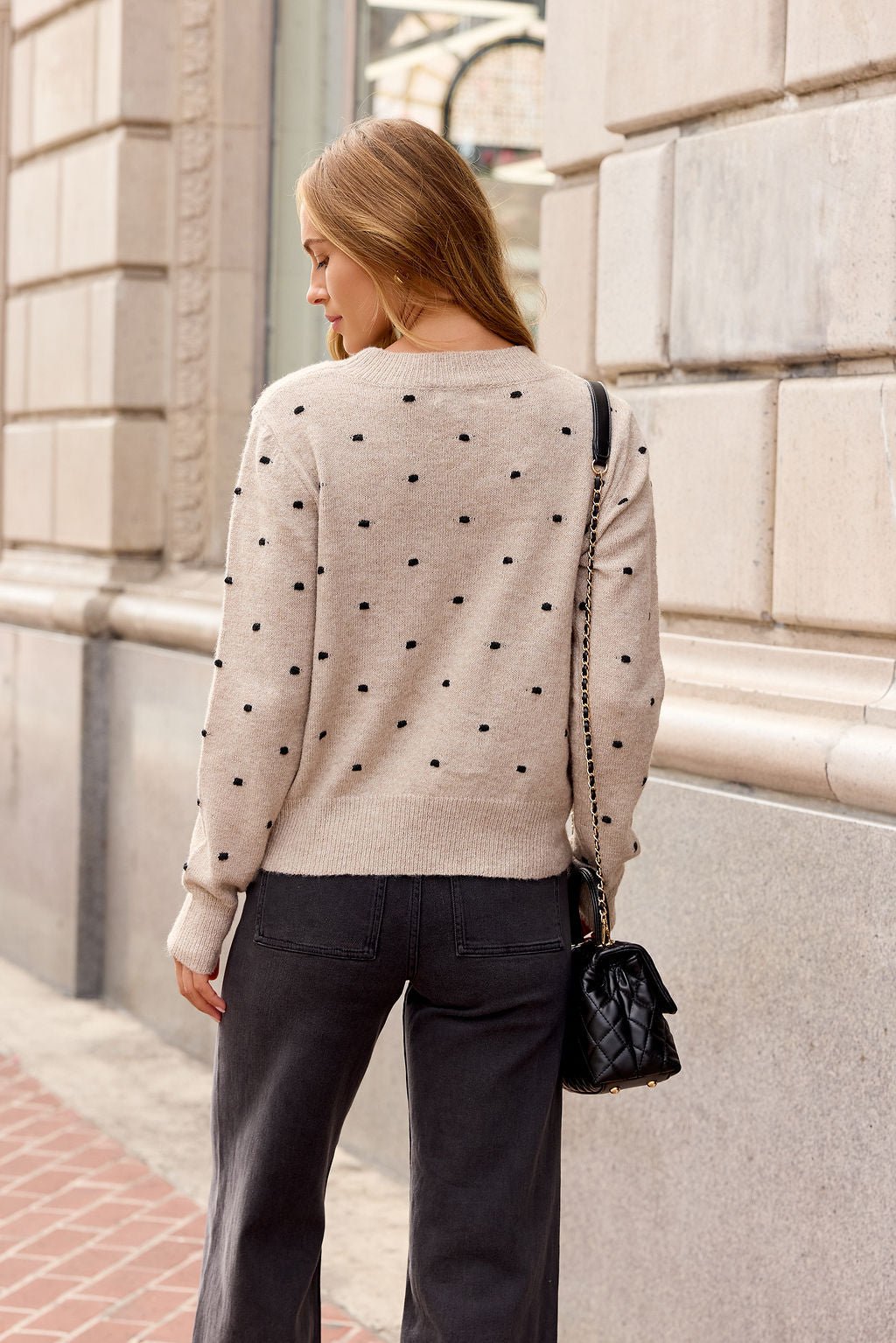 Woman with long hair in an Oatmeal Ribbed Polka Dot Sweater, black bag, and dark pants—perfect for fall—in the city.