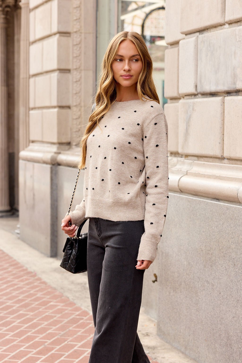 A woman in an Oatmeal Ribbed Polka Dot Sweater and dark pants stands on a city sidewalk, holding a black handbag.