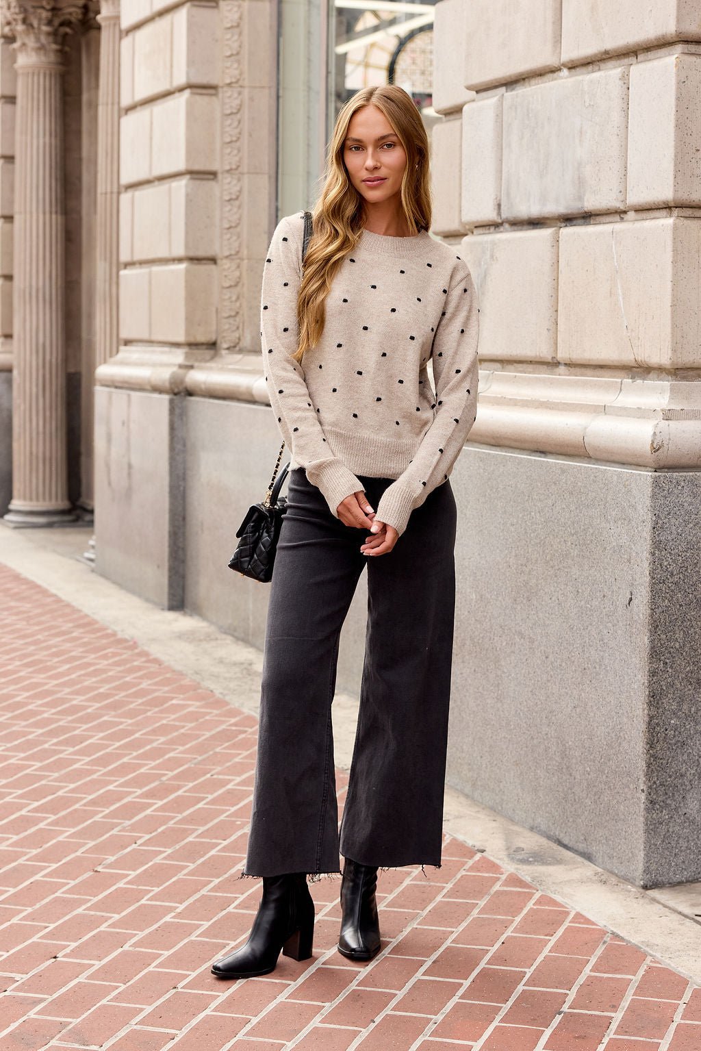 Woman in an Oatmeal Ribbed Polka Dot Sweater, black wide-leg pants, and boots stands on a brick sidewalk near a stone building.