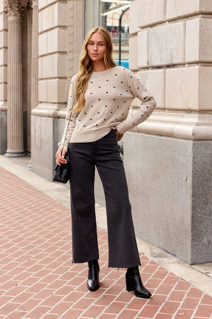 Woman in an Oatmeal Ribbed Polka Dot Sweater, black wide-leg jeans, and boots stands on a city sidewalk near a stone building.