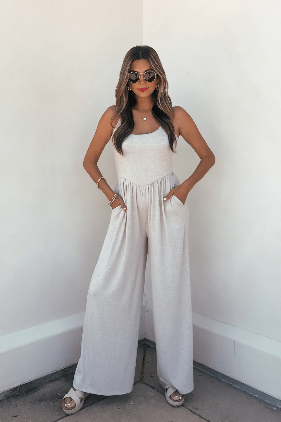 Woman wearing sunglasses and an Oatmeal Scoop Neck Wide Leg Jumpsuit, standing with hands in pockets against a white wall.