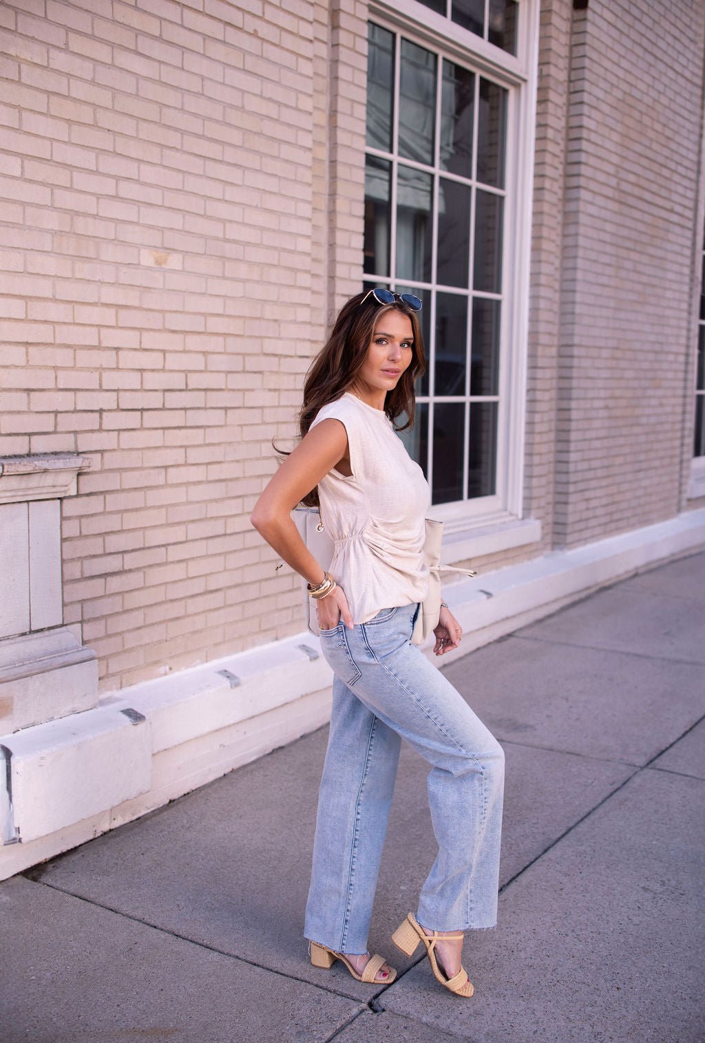 Woman in an Oatmeal Shirred Linen Top, jeans, and sandals stands on a city sidewalk near a brick building with large windows.