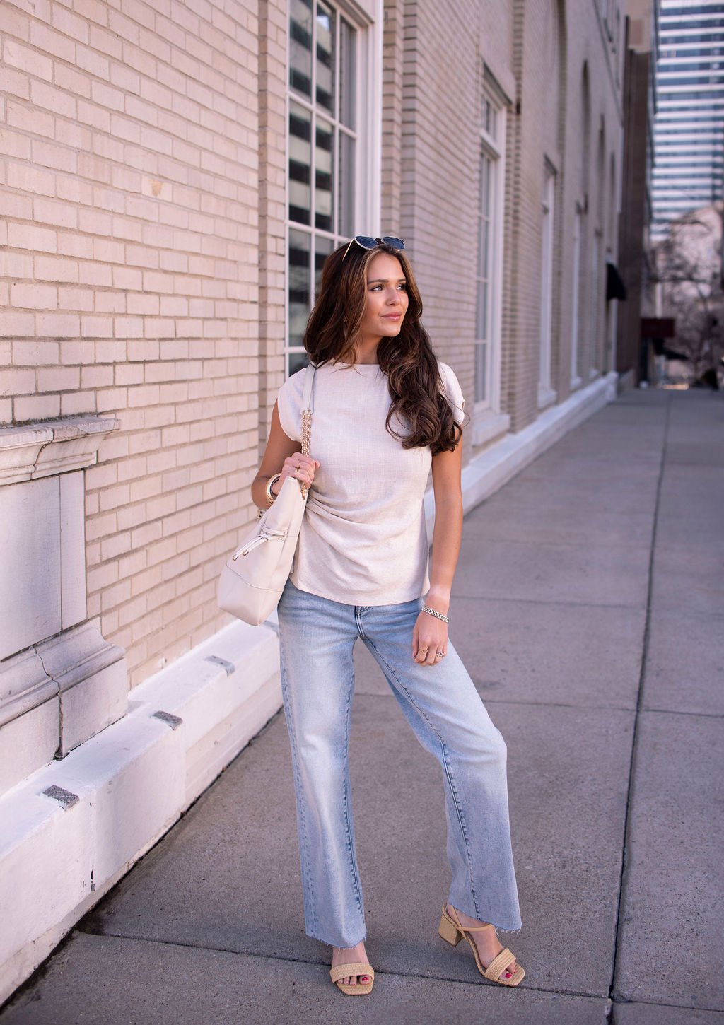 A woman in an Oatmeal Shirred Linen Top, light blue jeans, sandals, and a white bag stands on a city sidewalk.