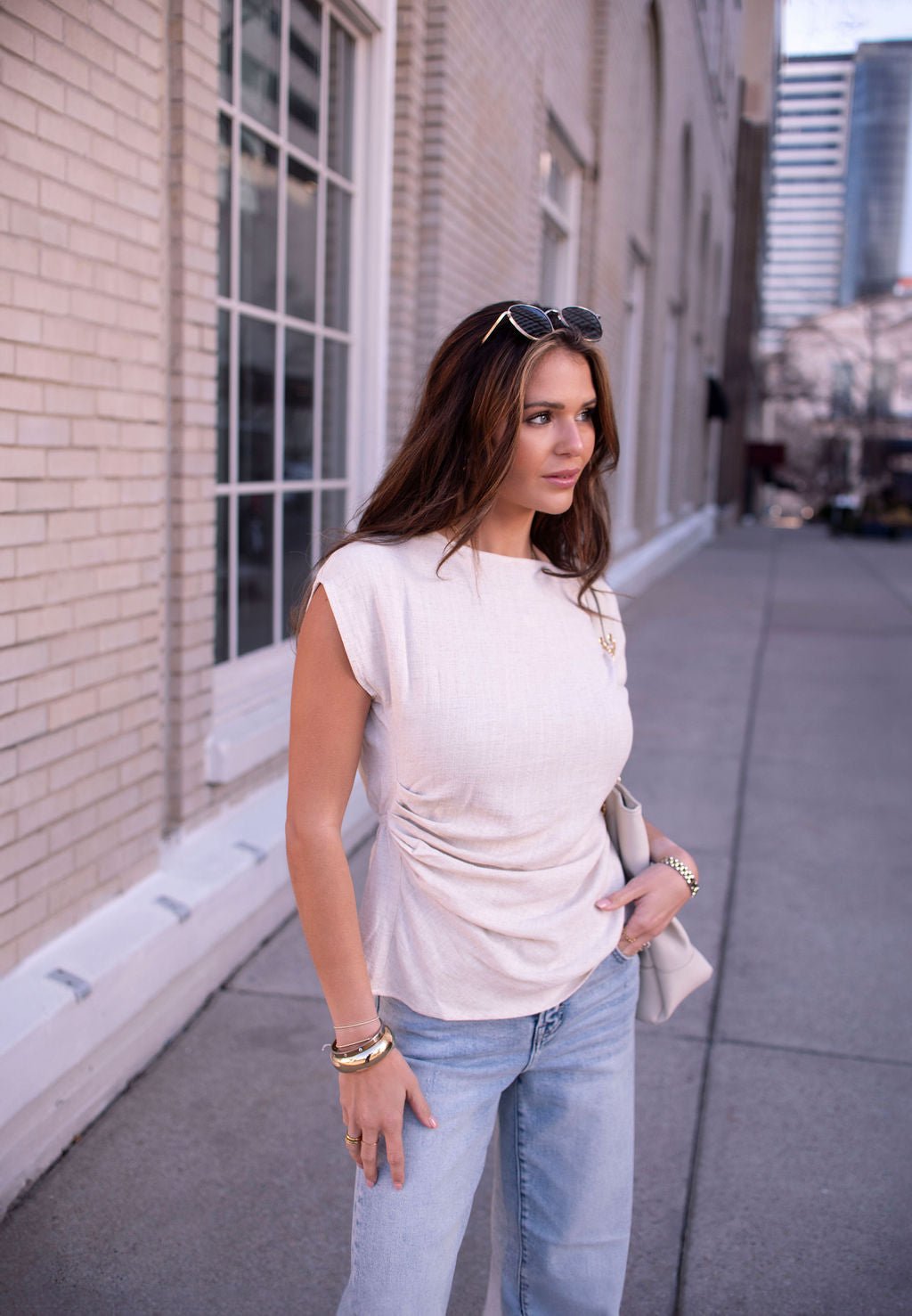 Woman in an Oatmeal Shirred Linen Top and light blue jeans stands on a city sidewalk, perfect for warm-weather styling.