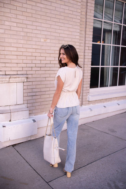 Woman wearing an Oatmeal Shirred Linen Top and light blue jeans smiles, walking on a sidewalk holding a large white handbag.