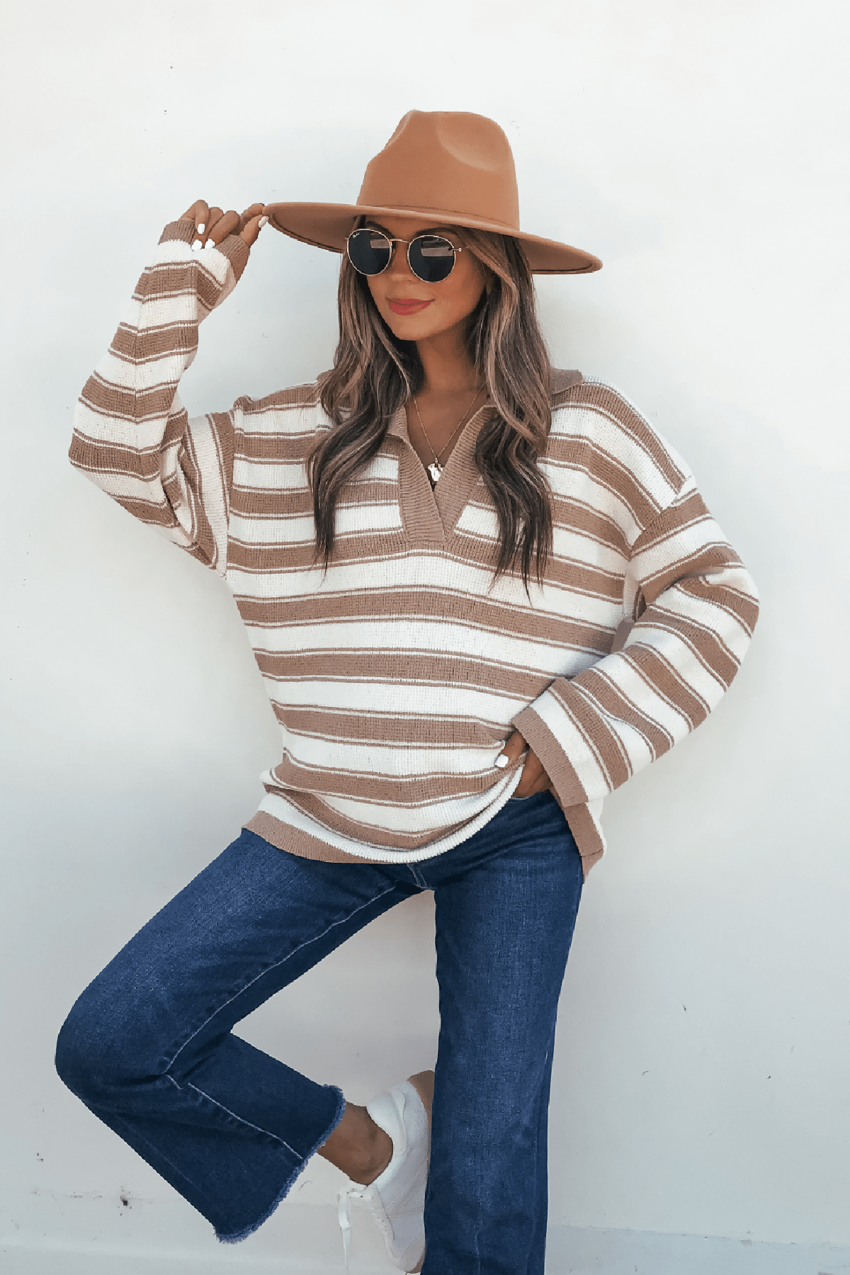 Woman in an Oatmeal Split V Neck Striped Sweater, jeans, tan hat, and sunglasses poses with one leg raised against a white wall.