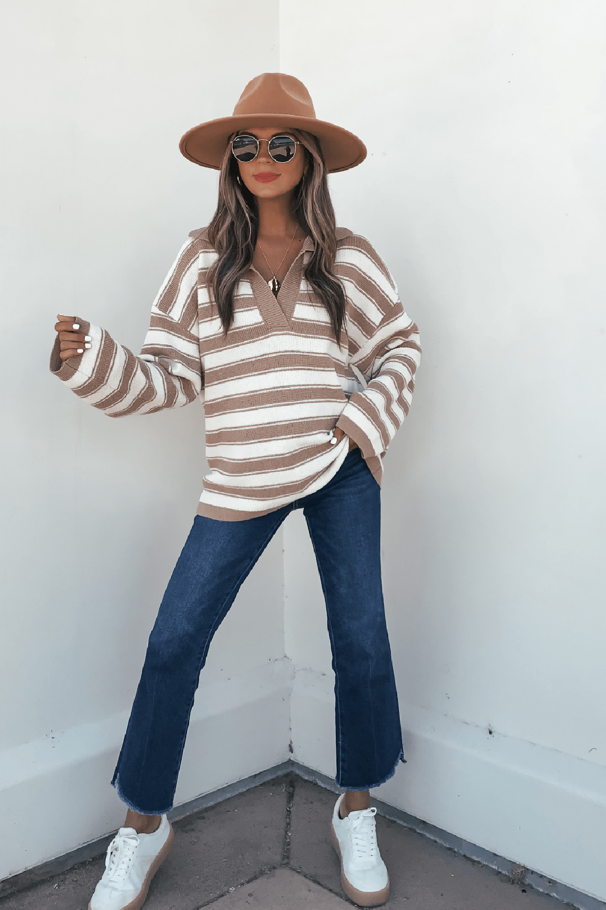 Woman in an Oatmeal Split V Neck Striped Sweater, tan hat, blue jeans, and white sneakers poses against a white wall.