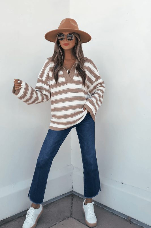 Woman in Oatmeal Split V Neck Striped Sweater, blue jeans, white sneakers, brown hat, and sunglasses against a white wall.