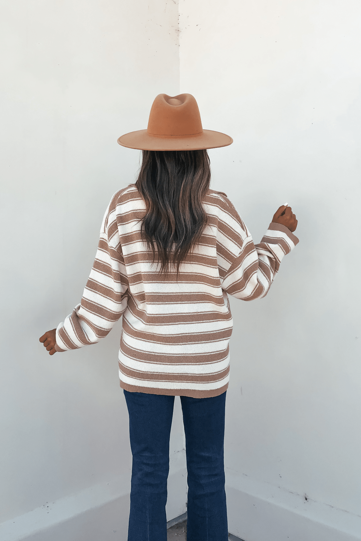 Woman in a brown hat, Oatmeal Split V Neck Striped Sweater, and blue jeans stands with her back to a white wall.