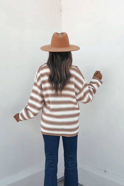 Woman in a brown hat, Oatmeal Split V Neck Striped Sweater, and blue jeans stands with her back to a white wall.