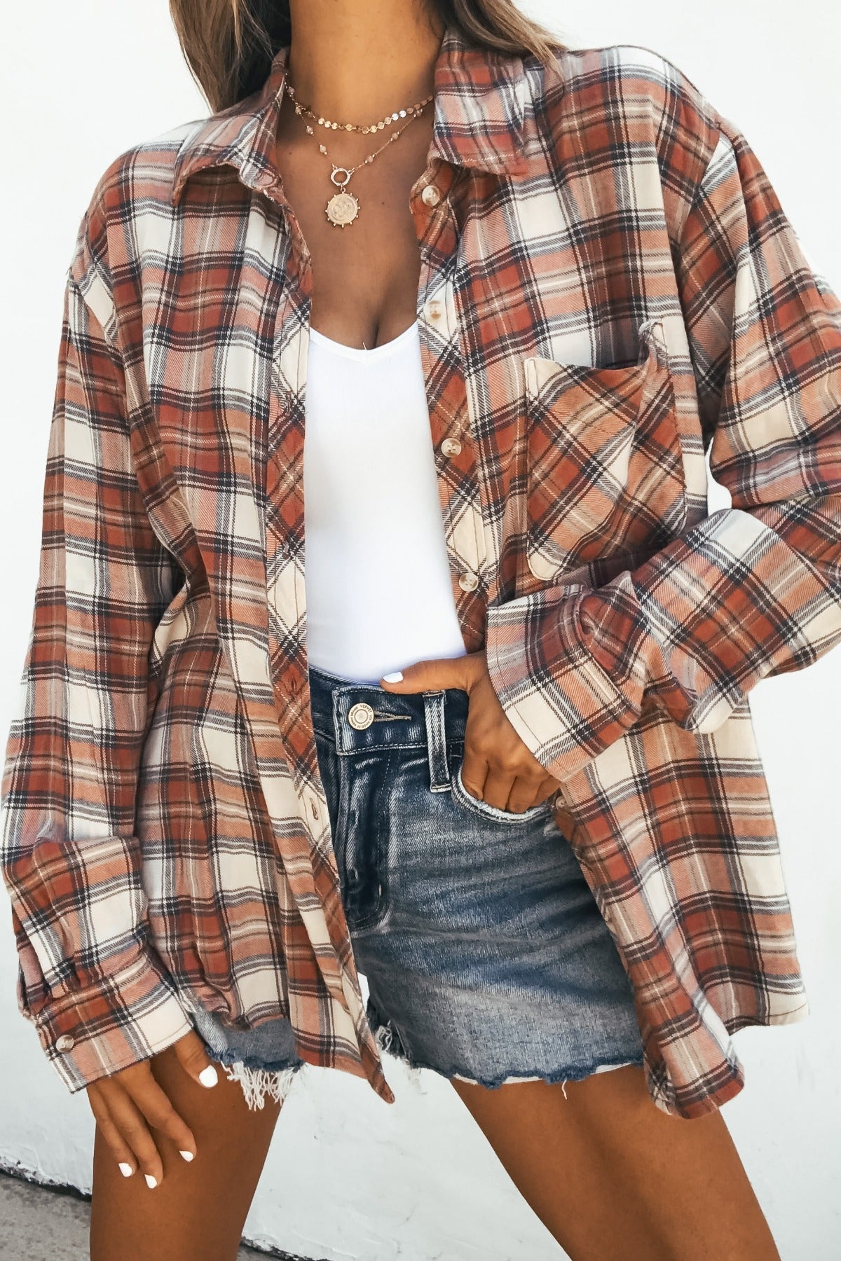 Model in Off The Grid Plaid Flannel Shirt - Red over a white tank and denim shorts, posing with hand on hip.