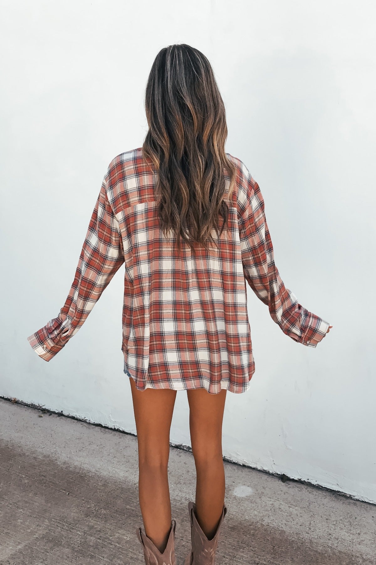 Woman standing outdoors in the Off The Grid Plaid Flannel Shirt - Red, arms outstretched, facing a white wall.