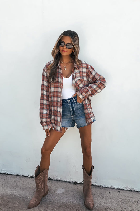 Woman in sunglasses, Off The Grid Plaid Flannel Shirt - Red, denim shorts, and cowboy boots poses confidently against a white wall.