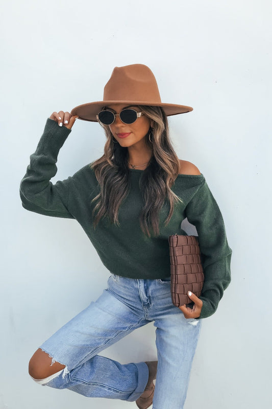 Woman wears an Off The Shoulder Sweater Top in dark green with ripped jeans, brown hat, and clutch against a white background.