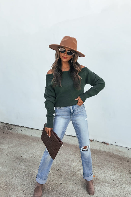 Woman in a brown hat, dark green Off The Shoulder Sweater Top, ripped jeans, and boots poses by a white wall holding a brown clutch.