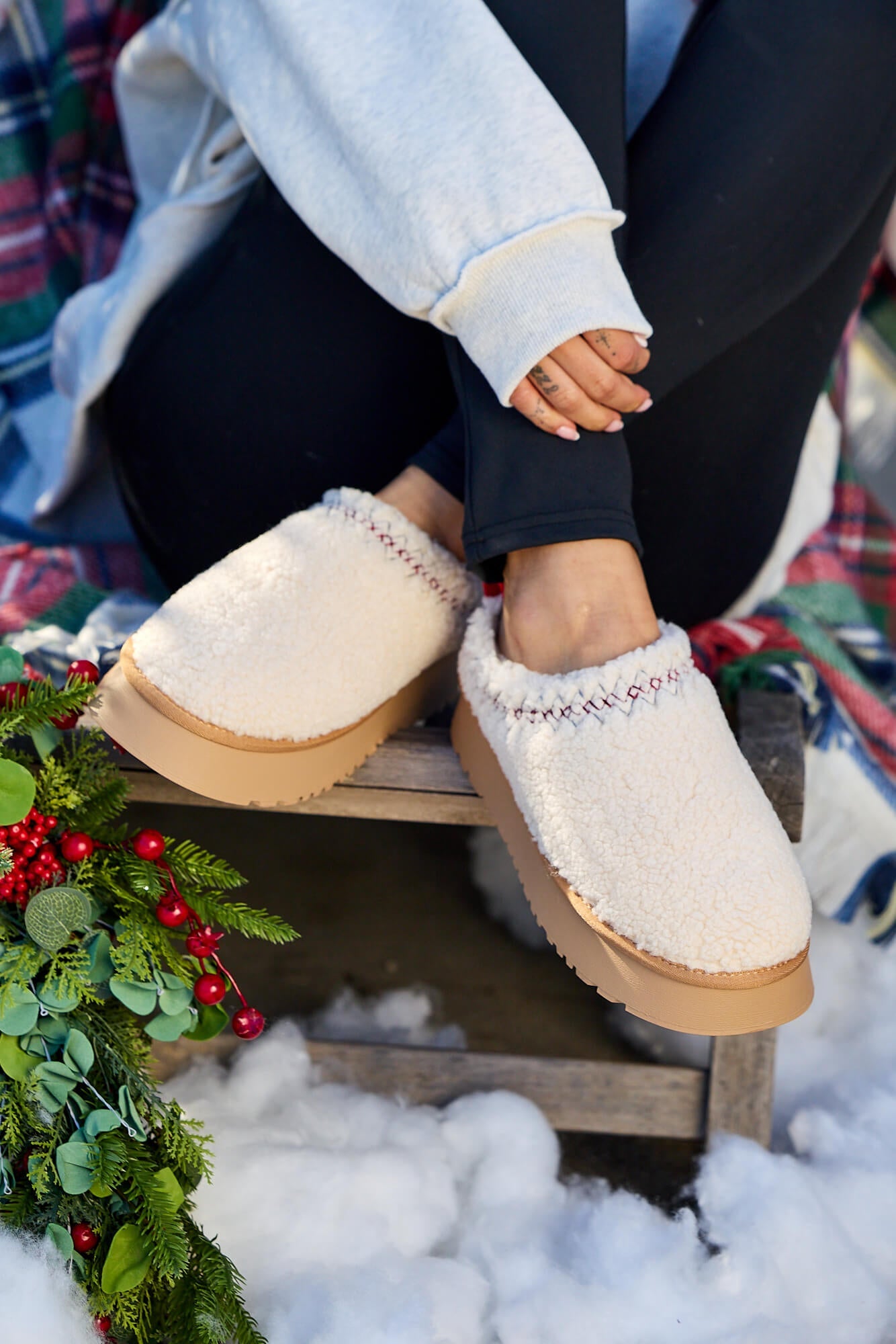 A person in Off White Sherpa Platform Slippers and black leggings sits on a bench amid snow, a plaid blanket, and holiday greenery.