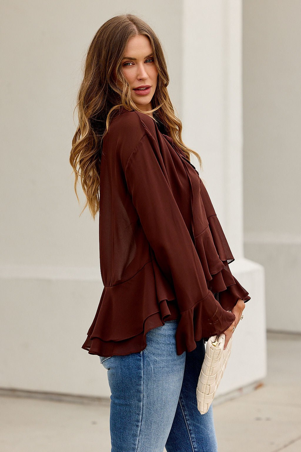 Woman with long wavy hair in the Office Bell Sleeve Top - Brown and blue jeans holds a white clutch outdoors by a light-colored wall.
