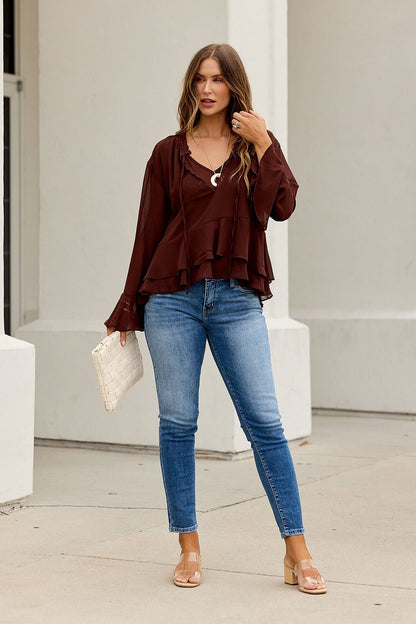 Woman in Office Bell Sleeve Top - Brown, blue jeans, and sandals holds a white clutch on a sidewalk by a white building.