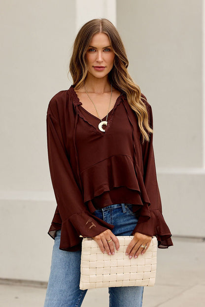 Woman with long wavy hair in Office Bell Sleeve Top - Brown and jeans holds a beige clutch against a light background.