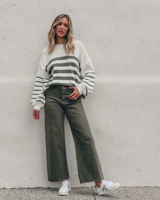 Woman in an Olive and Cream Striped Pullover Sweater, wide-leg olive pants, and white sneakers stands by a plain white wall.