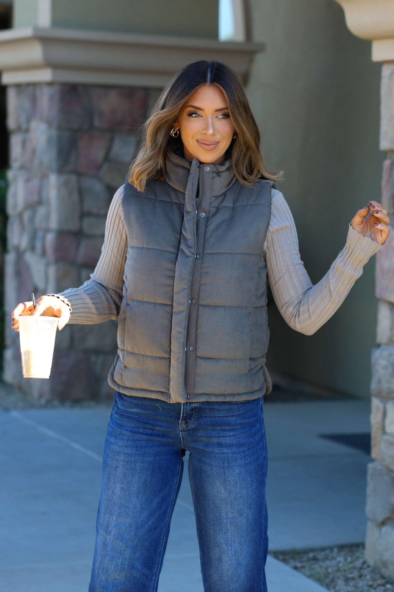 A woman in an Olive Button Up Puffer Vest smiles with a drink, showing off chic fall layering near a stone wall.