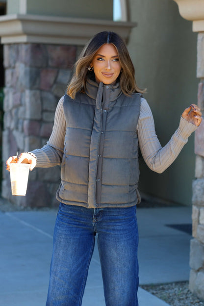 A woman in an Olive Button Up Puffer Vest smiles with a drink, showing off chic fall layering near a stone wall.