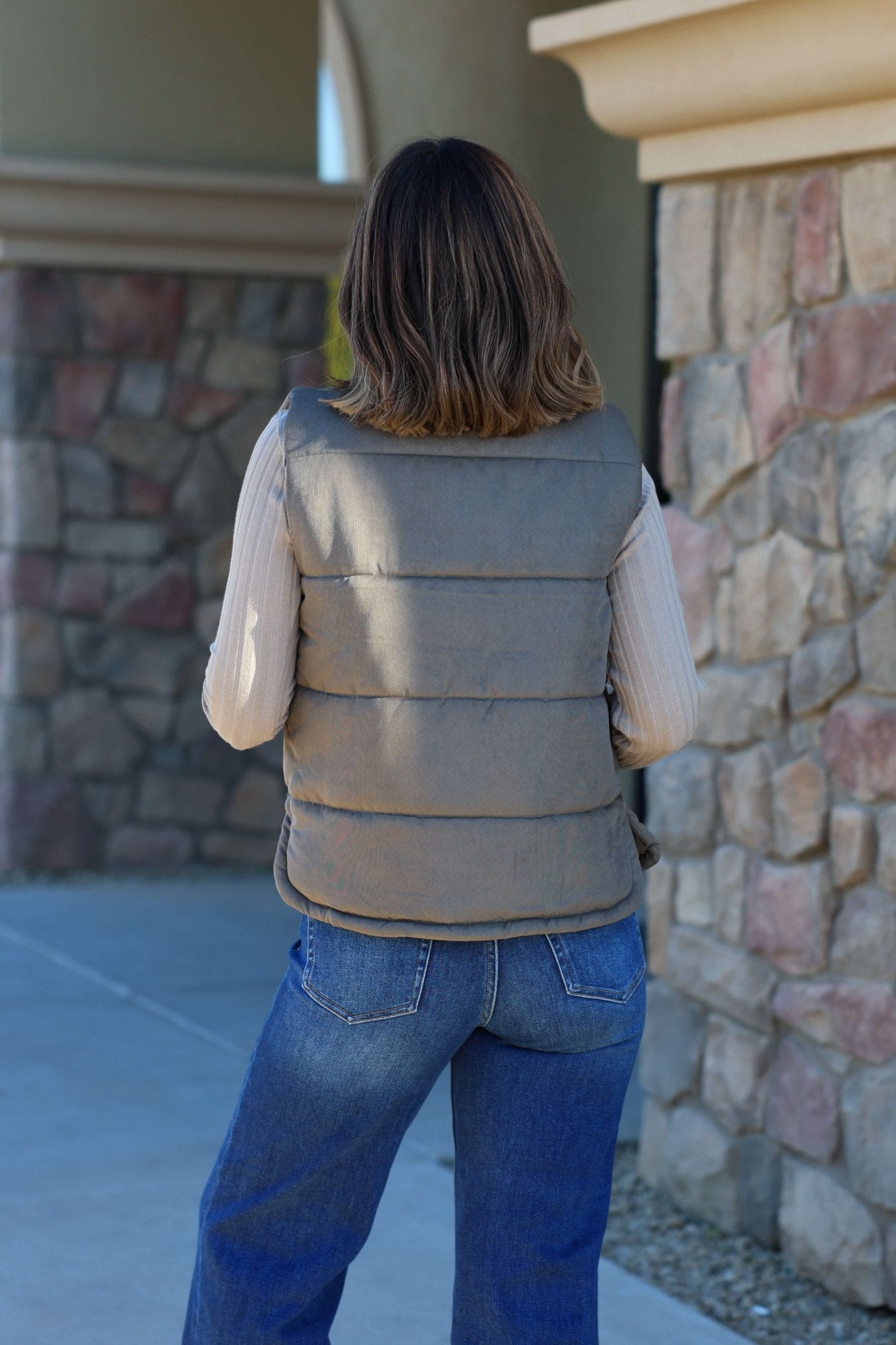 A woman in the Olive Button Up Puffer Vest and blue jeans stands by a stone wall—an ideal autumn look.
