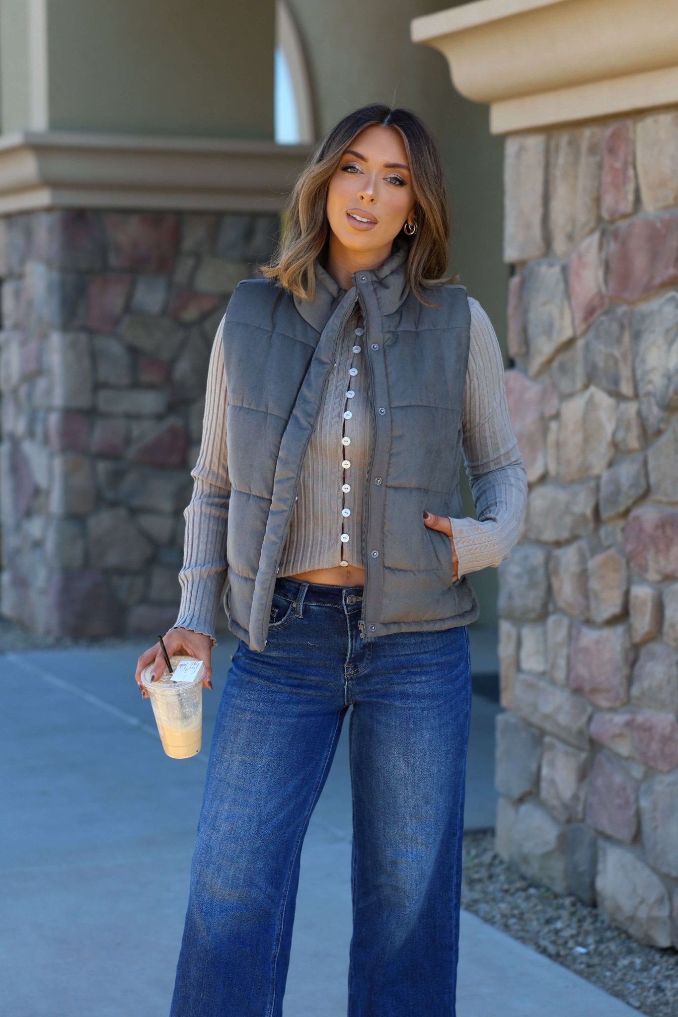 Woman outside in an olive button up puffer vest, ribbed sweater, wide-leg jeans, holding an iced coffee.