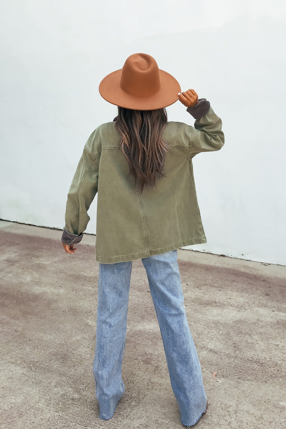 Person with long hair in a brown hat and blue jeans wears the Olive Contrast Collared Jacket, standing on concrete facing away.