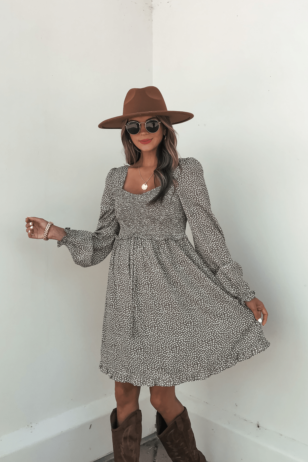 Woman wearing sunglasses, a brown hat, and the Olive Floral Print Ruched Mini Dress - FINAL SALE poses stylishly against a white wall.