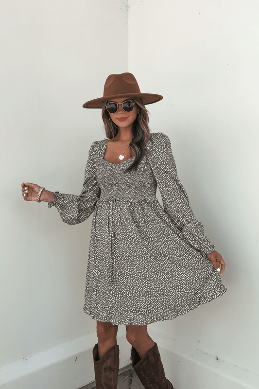 Woman poses in an Olive Floral Print Ruched Mini Dress - FINAL SALE, styled with a brown hat, sunglasses, and boots.