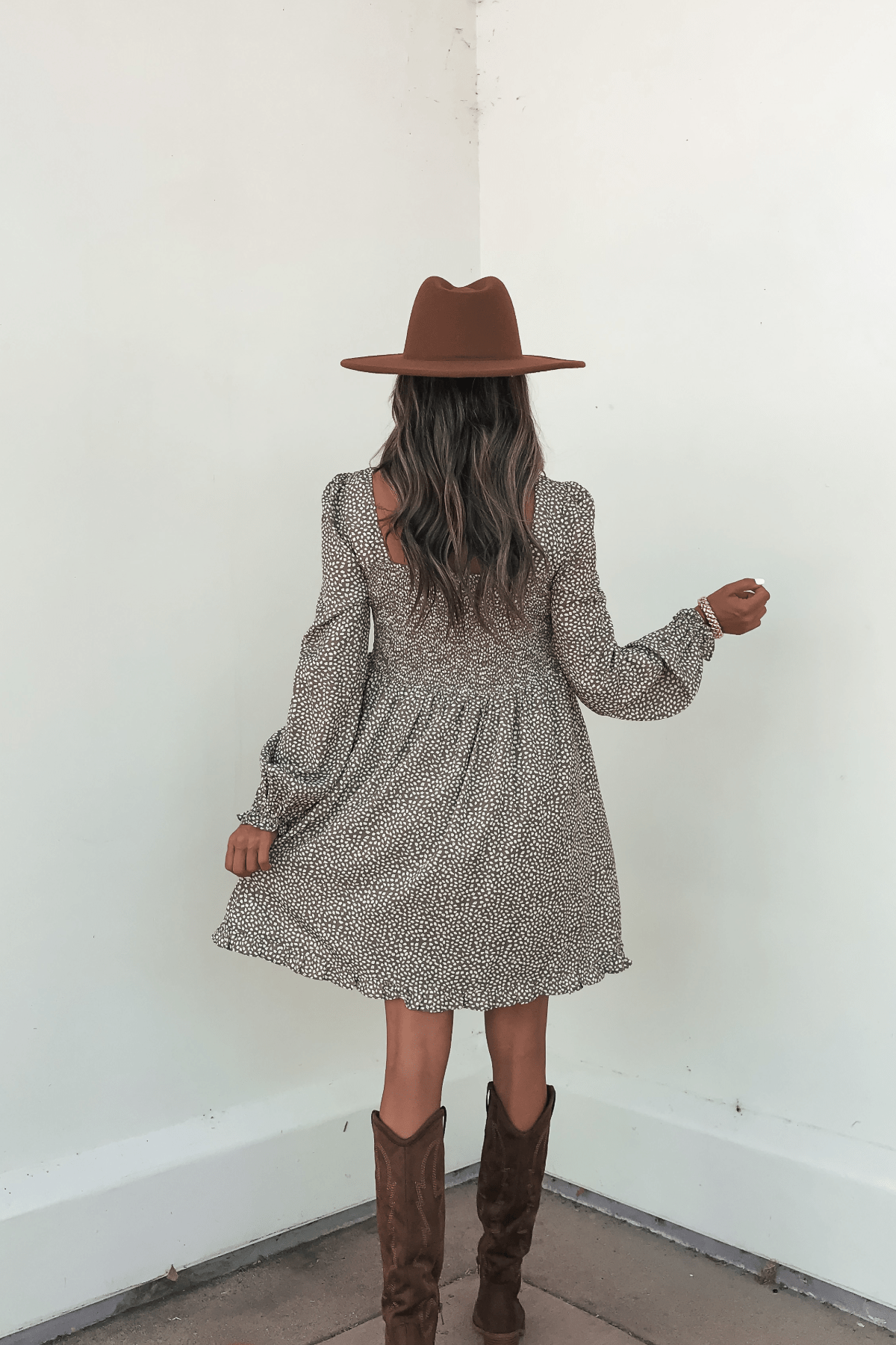 A woman in a brown hat and boots stands facing a white wall, holding her Olive Floral Print Ruched Mini Dress - FINAL SALE.